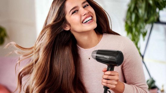 Vrolijke vrouw met lang bruin haar houdt een zwarte Philips föhn vast.