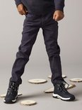 Jongensbenen in donkerblauwe broek met zwarte veterschoenen, omringd door houtsnippers.