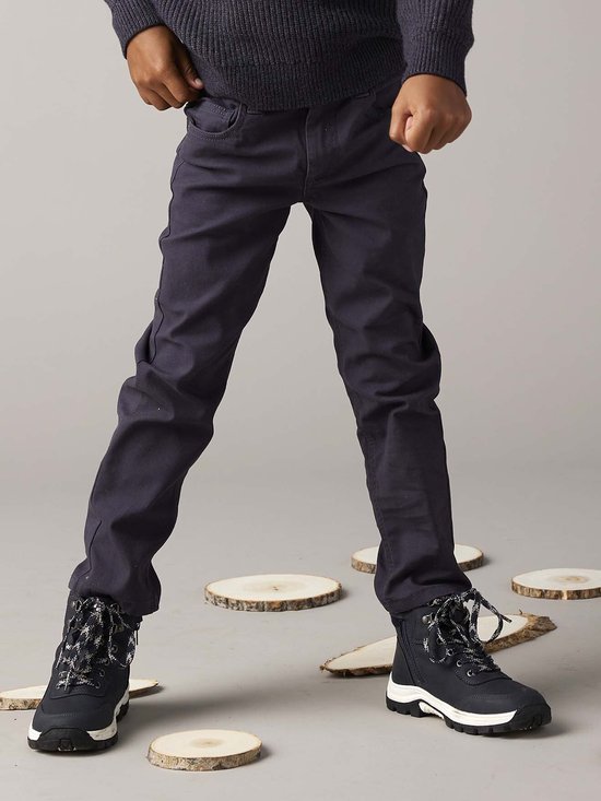Jongensbenen in donkerblauwe broek met zwarte veterschoenen, omringd door houtsnippers.