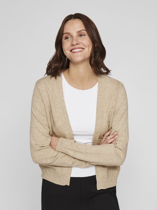 Een lachende vrouw met gekruiste armen, draagt een beige vest over een wit shirt.