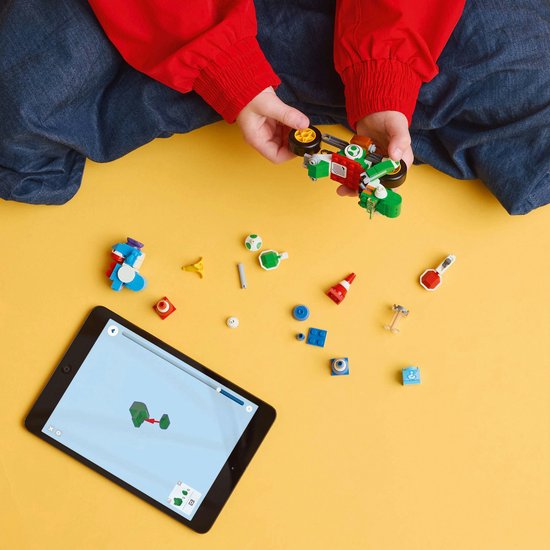 Handen van een kind bouwen een groene Lego-motor met een blauwe Yoshi en pionnen, naast een tablet met Lego-instructies.