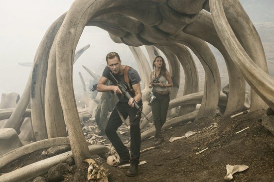 Tom Hiddleston en Brie Larson tussen gigantische botten, kijkend naar de camera in een mistig landschap.