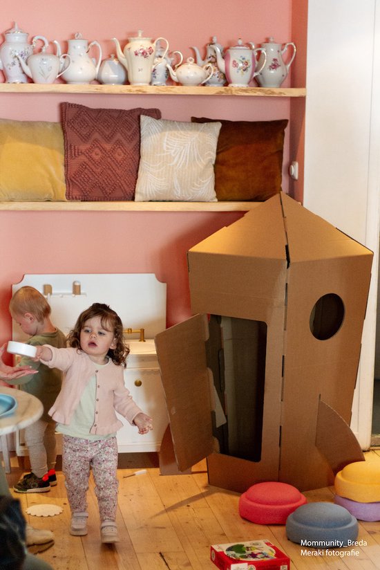 Grote kartonnen raket in een kamer met kinderen en decoratie. De deur staat open.