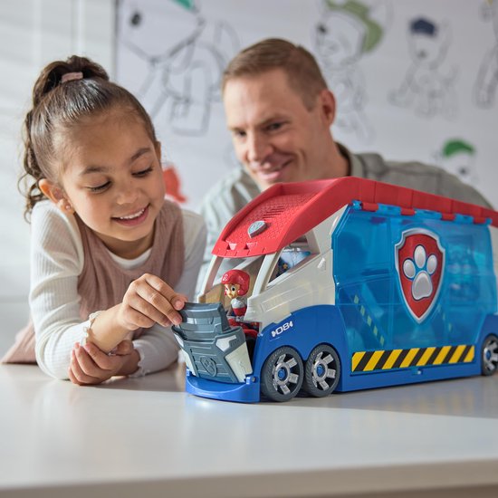 Een meisje speelt met de PAW Patrol Patroller, een groot blauw, wit en rood voertuig, met haar vader die toekijkt.