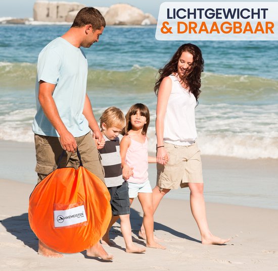 Gezin met twee kinderen loopt op het strand, vader draagt oranje pop-up tent in draagtas.