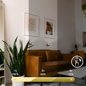 Zwarte wandlamp met verstelbare helderheid (100%, 50%, 15%) en warme verlichting, geplaatst in een woonkamer naast een bank.