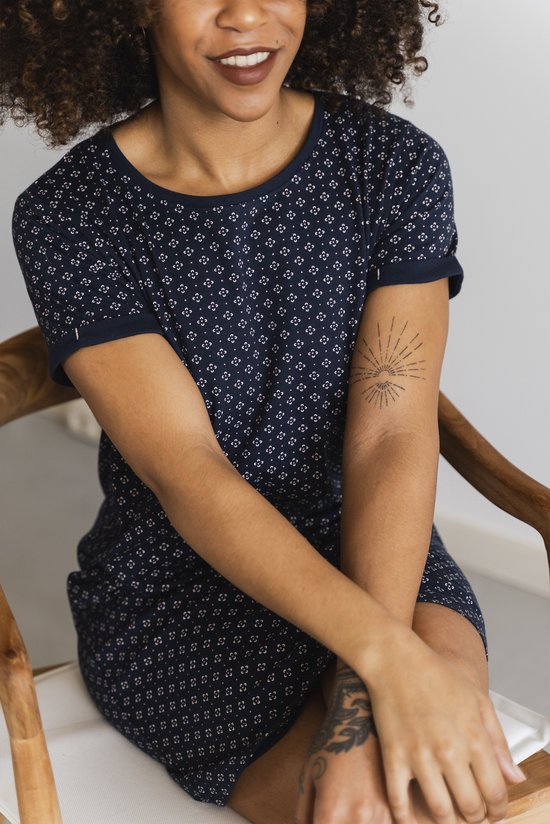 Zwarte vrouw zit op een stoel in een nachthemd met korte mouwen en een repetitief patroon van kleine witte bloemetjes.