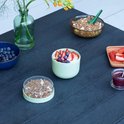 Een lichtgroene beker met yoghurt en fruit, omringd door muesli, blauwe bessen en aardbeien op een donkere tafel.
