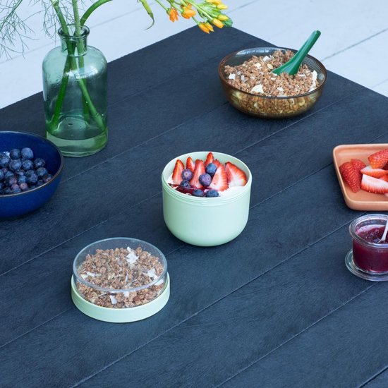 Een lichtgroene beker met yoghurt en fruit, omringd door muesli, blauwe bessen en aardbeien op een donkere tafel.