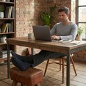 Man in grijze broek en grijs shirt werkt aan een laptop aan een houten tafel. Hij draagt Sukats antraciet sokken met rode streep.