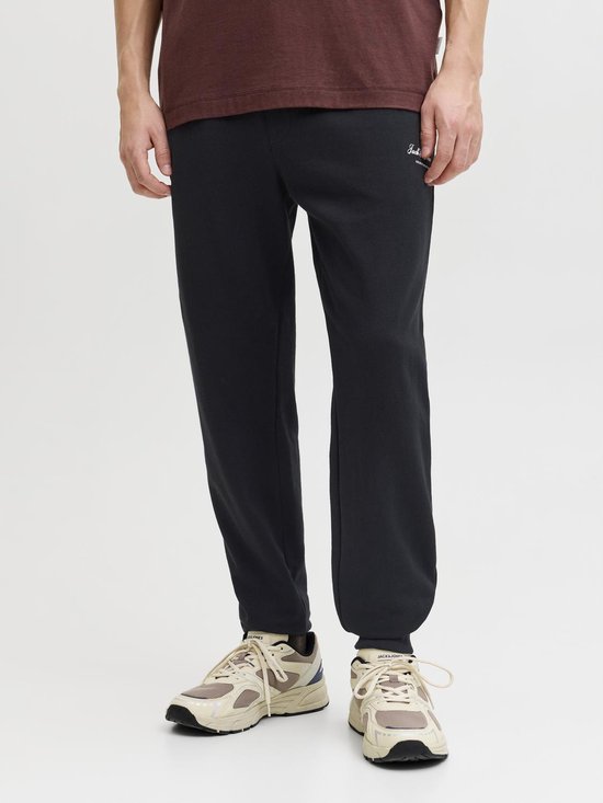 Man draagt een zwarte joggingbroek met witte 'Jack & Jones' opdruk, beige sneakers.