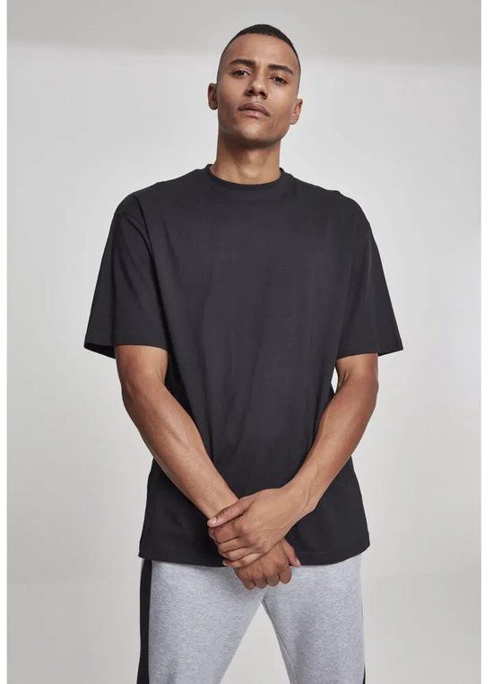 Man staat frontaal in zwart oversized T-shirt en grijze joggingbroek met zwarte streep, handen gevouwen.