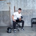 Jonge man met witte t-shirt en zwarte trainingsbroek zit op een bankje en tilt een dumbbell.