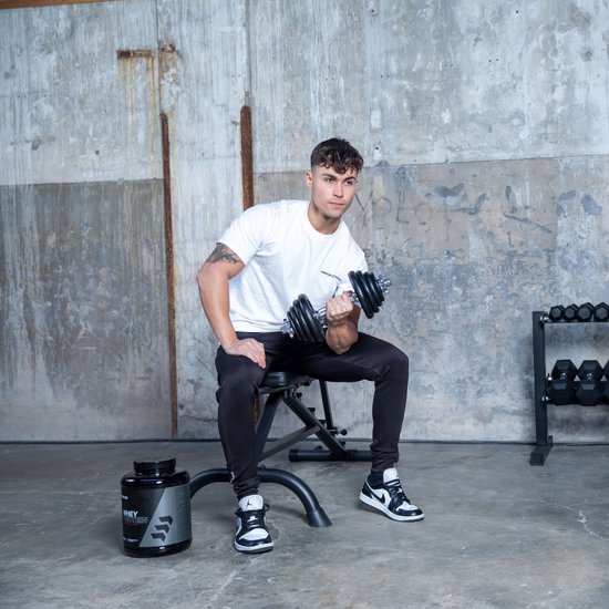 Jonge man met witte t-shirt en zwarte trainingsbroek zit op een bankje en tilt een dumbbell.