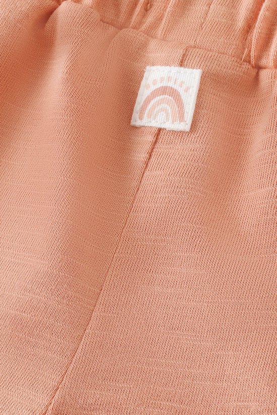 Koraalkleurige baby short met elastische tailleband en een label met een regenboog-icoon.