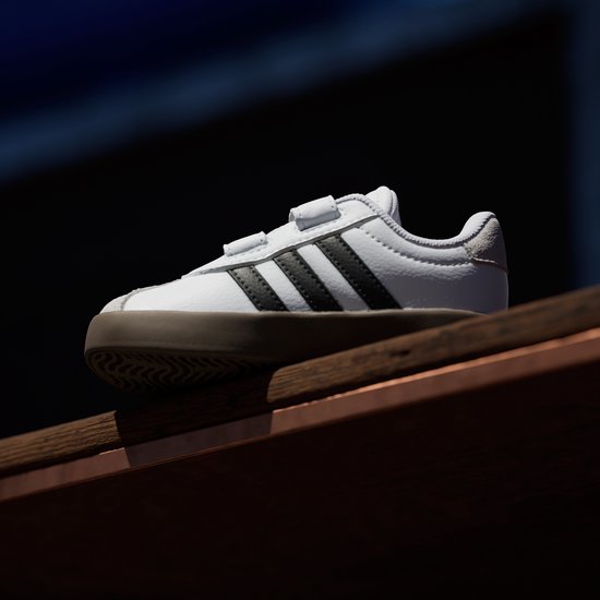 Witte sneaker met zwarte strepen en bruine zool, met klittenbandsluiting en grijze suède details.