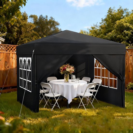 Zwarte partytent in een tuin, ingericht met een ronde tafel gedekt voor een maaltijd en witte stoelen.