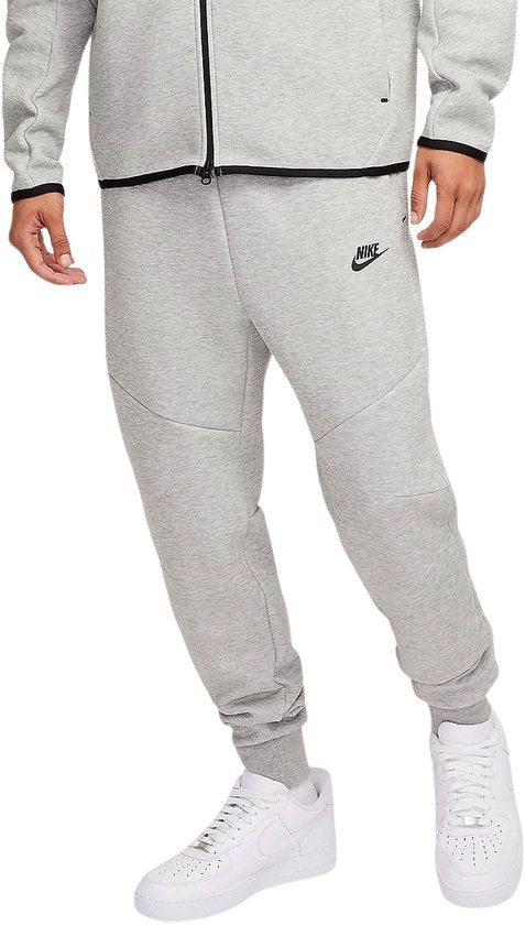 Man in een grijze Nike joggingbroek en wit Nike logo, met ritssluiting op de zijkant.