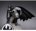 Batman-figuur in grijs pak met gele riem en zwarte cape, zittend op een lamp met vleermuislogo.