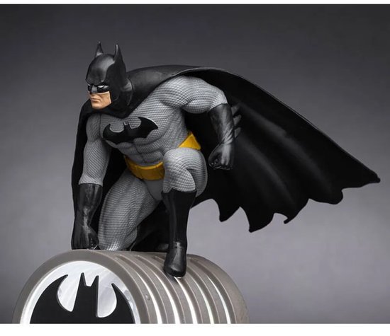 Batman-figuur in grijs pak met gele riem en zwarte cape, zittend op een lamp met vleermuislogo.