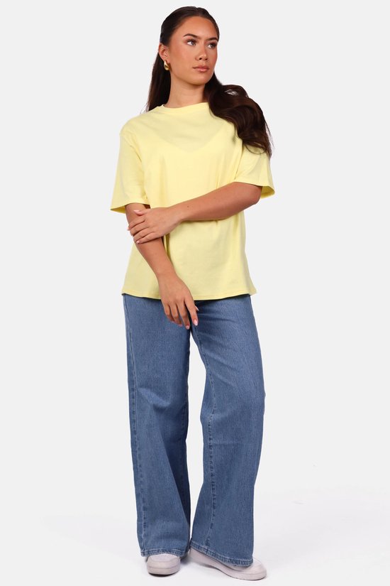 Model met gekruiste armen draagt een lichtgele oversized T-shirt met een blauwe spijkerbroek.
