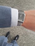 Bras portant une montre argentée avec bracelet métallique, cadran noir, heure analogique et numérique, avec un blazer bleu à carreaux.