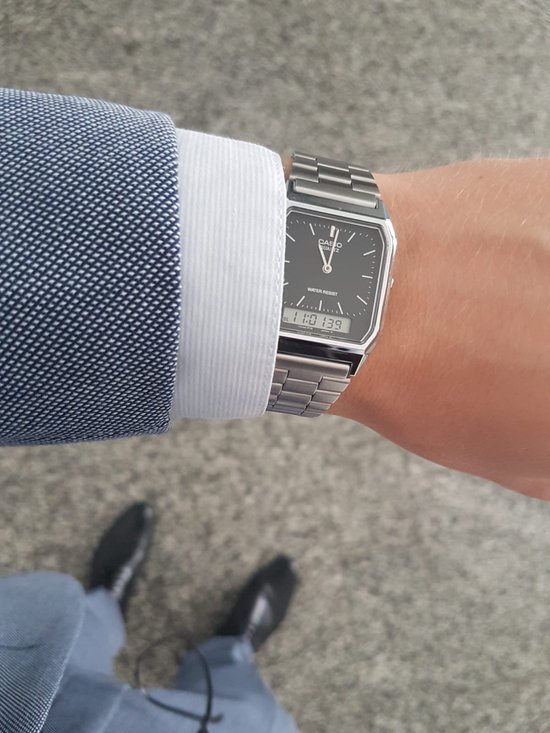Bras portant une montre argentée avec bracelet métallique, cadran noir, heure analogique et numérique, avec un blazer bleu à carreaux.
