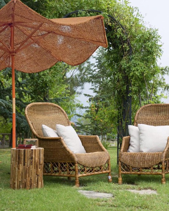 Bruine kokos parasol met golvende randen naast rieten stoelen in een tuin, met een bijzettafel en beplanting.