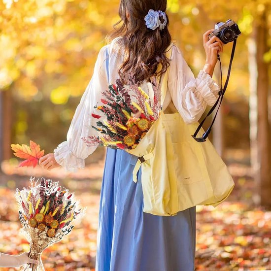 Vrouw met bruine krullen en blauwe bloem, draagt een geel tas met een boeket gedroogde bloemen, houdt een herfstblad vast.