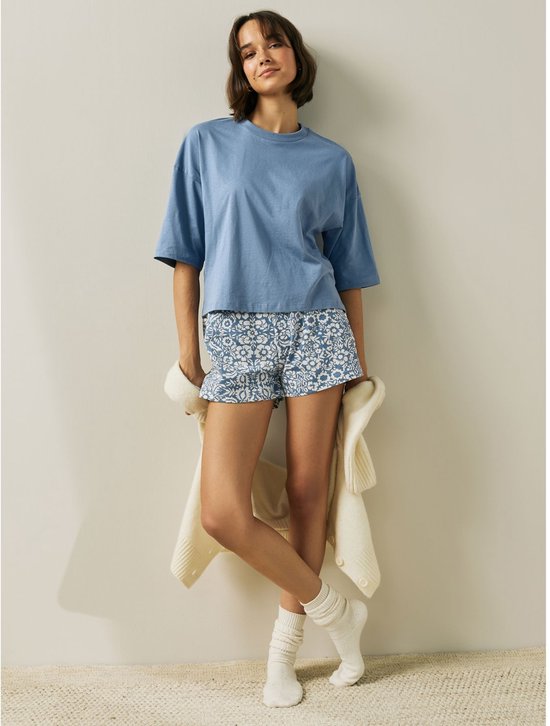 Model met blauw shirt en korte pyjamabroek met bloemenprint, houdt vest vast.