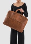 Een vrouw in een zwarte outfit draagt een bruine leren laptoptas met veel vakken en een schouderband.