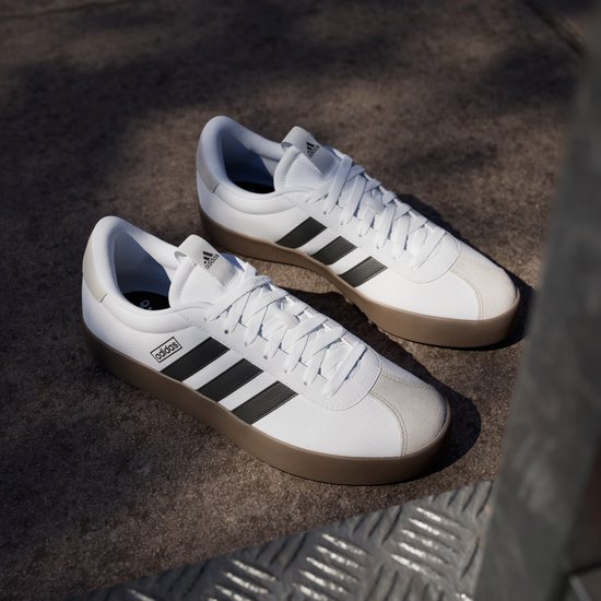Twee witte adidas sneakers met bruine zolen en zwarte strepen op een betonnen ondergrond.