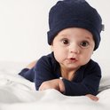 Baby met grote bruine ogen, liggend op de buik, draagt een donkerblauw boxpak en muts.