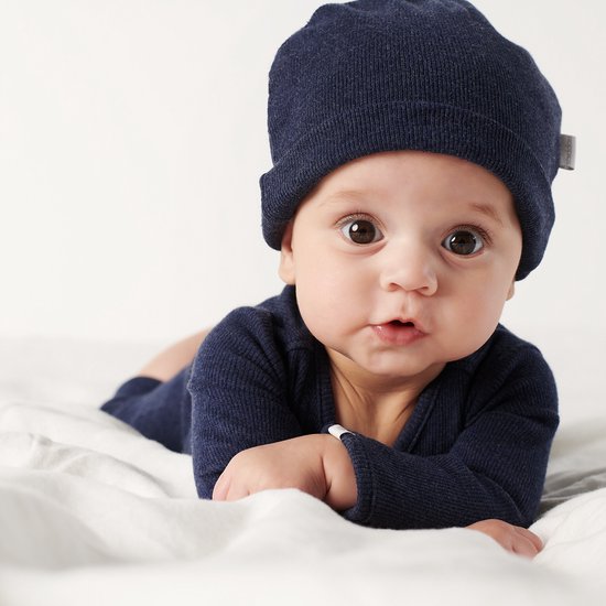 Baby met grote bruine ogen, liggend op de buik, draagt een donkerblauw boxpak en muts.