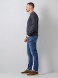 Man in een donkergrijs shirt en blauwe jeans, staand in profiel. Hij draagt bruine schoenen.