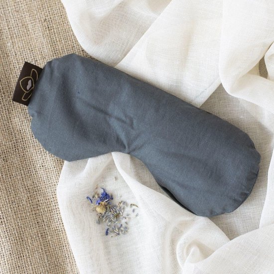 Grijs oogkussen met bruin label met lotuslogo, liggend op linnen stof met gedroogde bloemen.