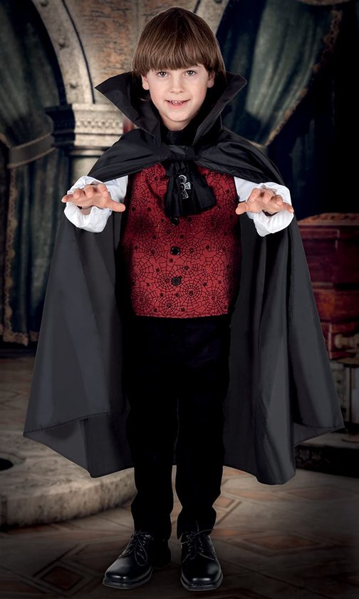 Jongen in vampierkostuum met zwarte cape, rood vest met spinnenwebben, wit shirt, zwarte broek, staand in een donkere kamer.