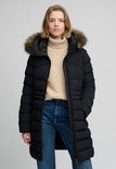 Lange zwarte pufferjas met bontkraag, gedragen door een blond model met beige coltrui en spijkerbroek.