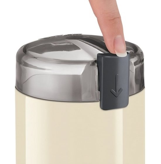 Een duim drukt op de grijze sluiting met naar beneden gerichte pijl, op een beige en metalen koffiemolen.