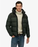 Man met beige coltrui en donkergroene, gewatteerde jas met capuchon, staand.
