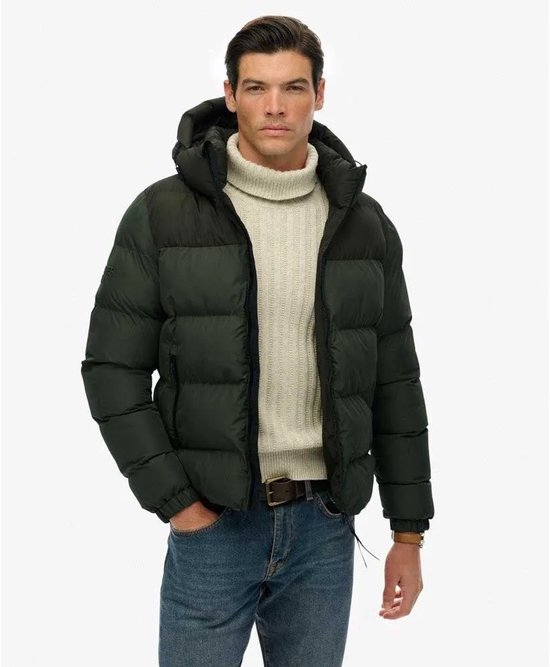 Man met beige coltrui en donkergroene, gewatteerde jas met capuchon, staand.