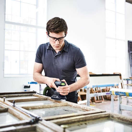Een man met veiligheidsbril schuurt een houten kozijn met een zwarte schuurmachine met groen accent.