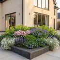 Moderne tuin met een bloembak gevuld met verschillende planten, waaronder paarse ooievaarsbekken, roze campanula's en witte bloemen.