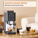 Grijze Krups volautomatische espressomachine op tafel, met glazen mokken koffie en thee.