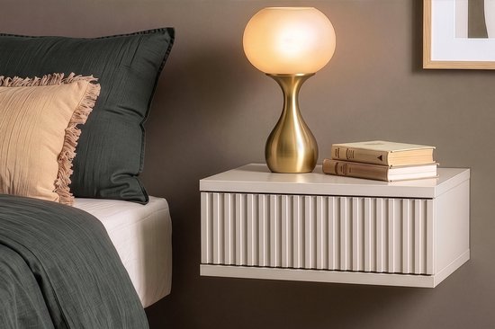 Beige hangend nachtkastje met verticale ribbels op ladefront, naast een bed met kussens en een lamp.