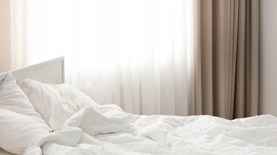 Opgebed wit bed bij een raam met taupekleurige gordijnen en witte vitrage.