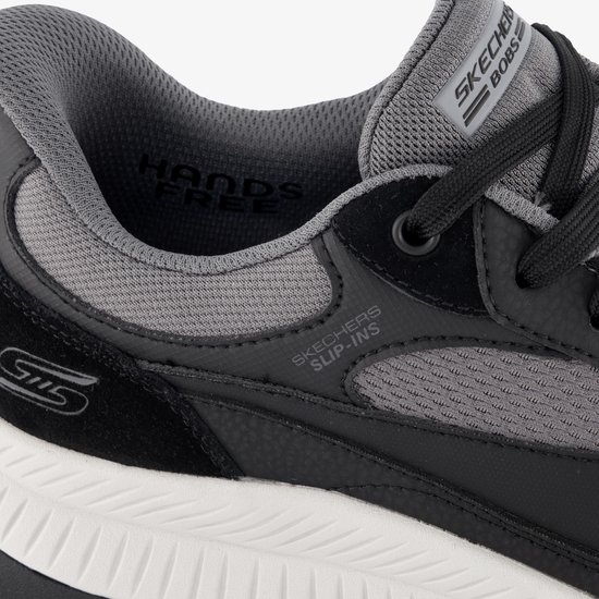 Detail van een zwarte sneaker met grijs mesh en S logo. Tekst 'HANDS FREE' en 'SKECHERS SLIP-INS' zichtbaar.