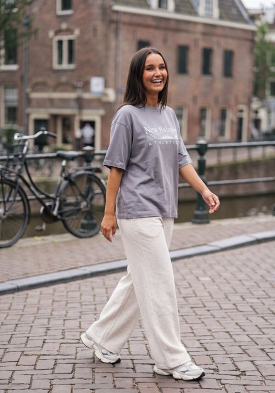 Vrouw in een grijze oversized New Balance T-shirt met atletische opdruk en een lichte beige broek, lopend over straat.
