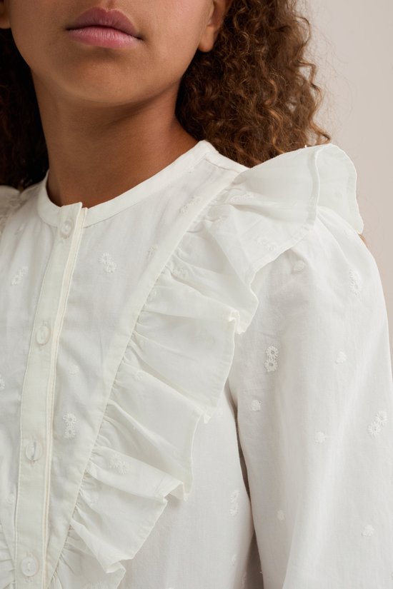 Close-up van een meisje met krullend haar, dat een witte blouse met ruches en geborduurde bloemetjes draagt.