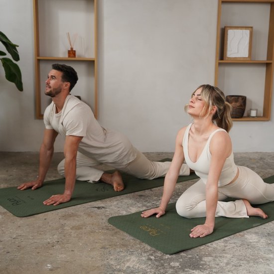 Man en vrouw in beige outfits doen yoga op donkergroene matten met gouden plantenprint. De pose is een cobra.